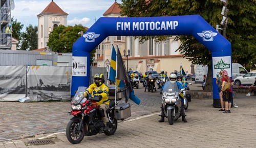 I förgrunden finns två motorcyklister som bär gula och blå kläder. Den ena motorcyklisten kör en röd motorcykel medan den andra kör en grå motorcykel. Båda motorcyklisterna bär hjälmar och verkar vara redo att köra.  I bakgrunden finns flera andra motorcyklister och en stor blå båge med texten "FIM MOTOCAMP". Det finns också ett blått flagg med FIM:s logotyp. Till höger i bilden står två personer bredvid en stolpe med flera skyltar. I bakgrunden finns byggnader, träd och en kyrkas torn.  Det verkar vara en samlingsplats eller startpunkt för ett motorcykelevenemang. Atmosfären ser festlig och organiserad ut med deltagare och åskådare som samlas för evenemanget.